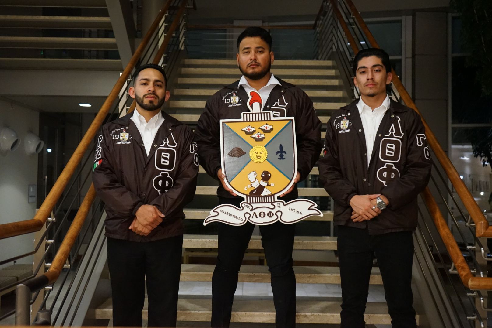 Three brothers in fraternity jackets on a staircase, the centre one holding a large wooden crest.