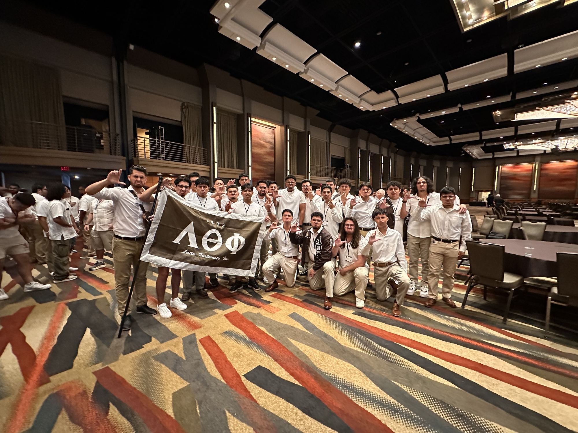 A large group of brothers in white shirts holding the Lambda Theta Phi flag in a hotel ballroom.