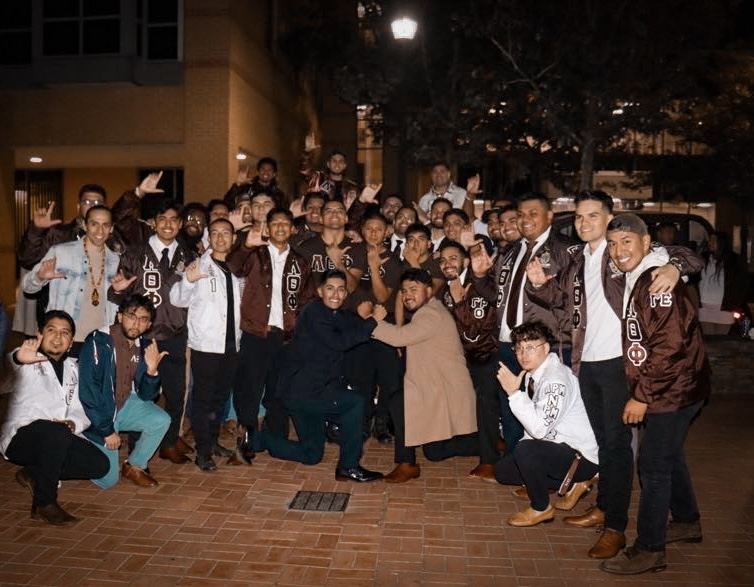 A large group of brothers outdoors at night on a brick walkway, throwing the hand sign.