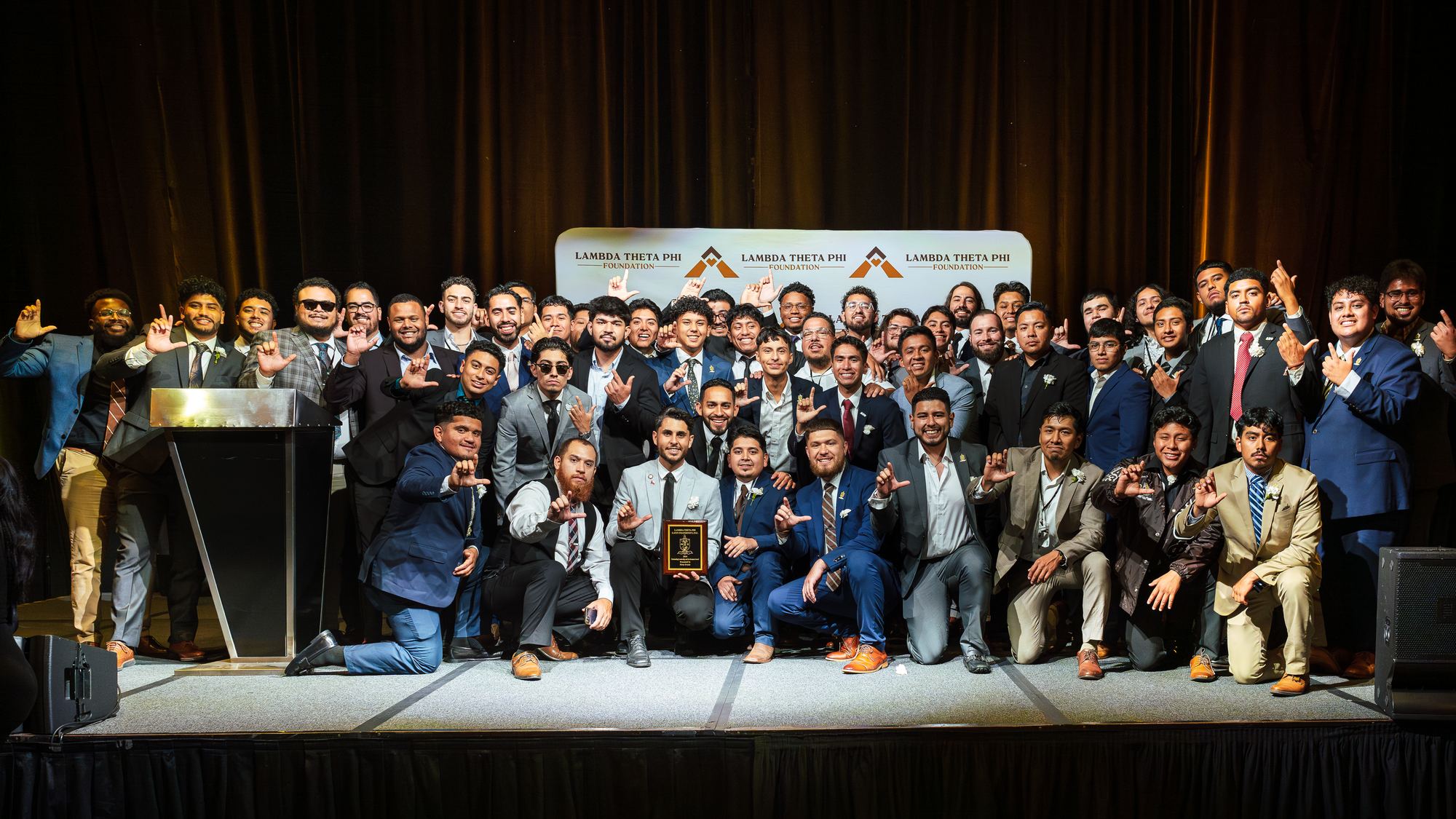 A large group of brothers on stage at a Lambda Theta Phi Foundation banquet, one holding an award plaque.