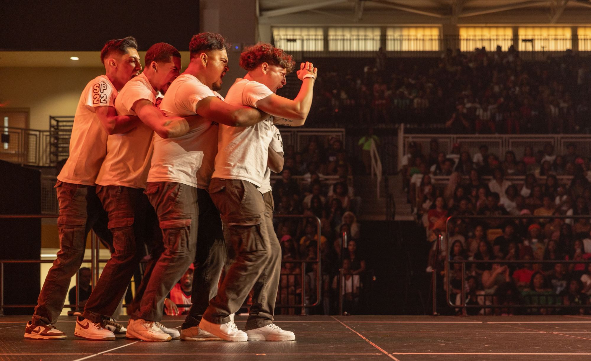 Four brothers strolling in a tight line on stage under red light, the arena crowd behind them.
